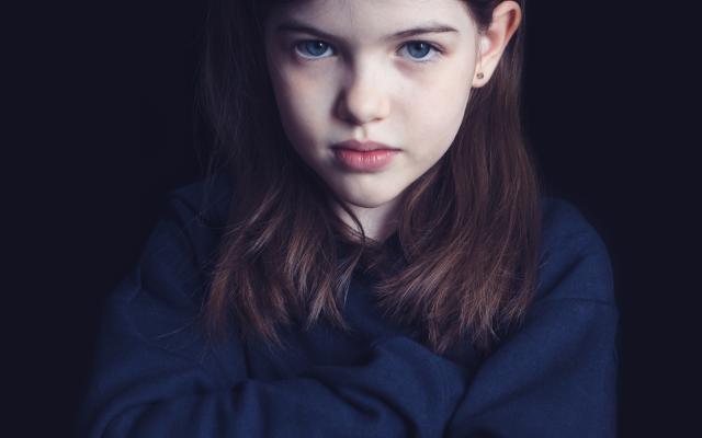Een jong meisje heeft haar armen over elkaar geslagen. Ze heeft een norse blik en kijkt recht in de camera.