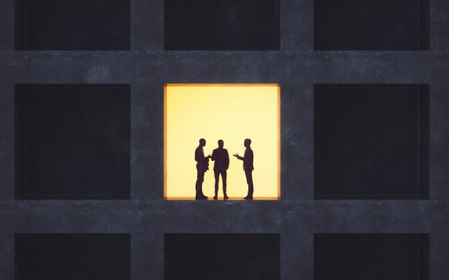 Drie silhouetten in gesprek in een verlicht raam van een verder donker gebouw.