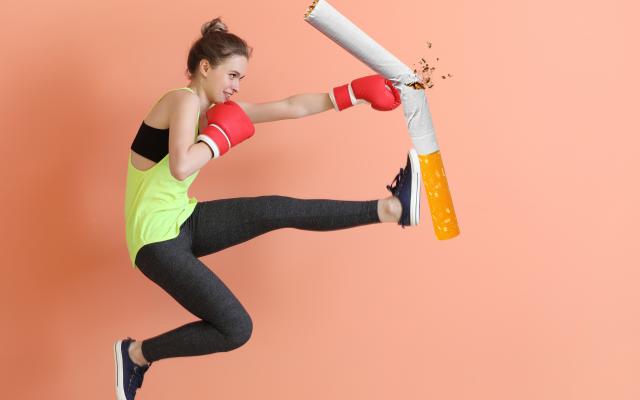 Een jonge vrouw draagt sportkleding en bokshandschoenen. Ze schopt en slaat tegen een grote sigaret.