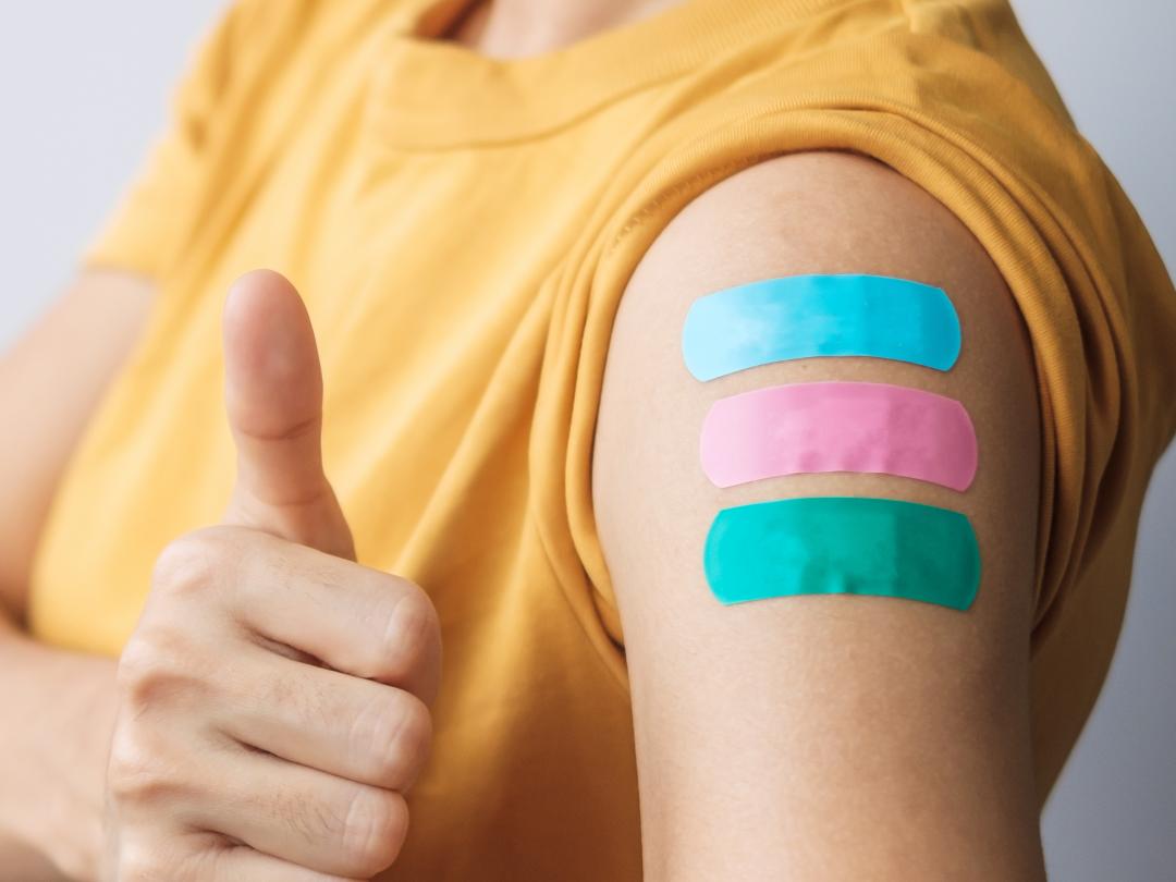 Een persoon met een mondkapje op en drie pleisters op de arm.