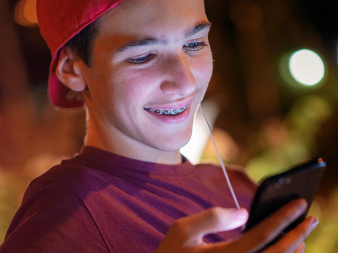 Een tiener met een beugel en een pet op kijkt lachend naar de telefoon in zijn hand.